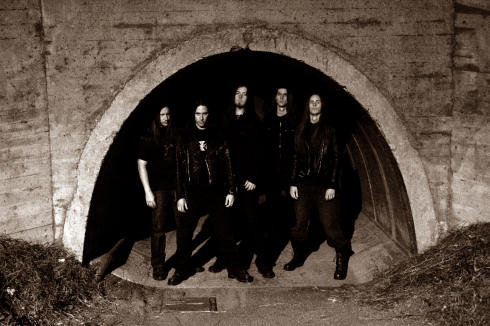 Viisi mustiin vaatteisiin pukeutunutta ja vakavailmeisestä Aeon-bändin miestä seisoo rivissä holvikaaren alla. Kyseessä on ulkoilmassa suuren betonisen holvikaarimaisen suuaukon edestä otettu kuva.