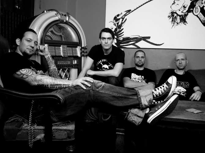 Harmaasävyinen Nikolaj Palmskovin ottama valokuva Volbeat-nimisestä metallibändistä, johon kuuluvat neljä miestä istuvat jukeboksin vieressä sohvilla ja tuoleilla. Heistä jokaisella lyhyet hiukset, mustat t-paidat yllä ja tyynet ilmeet kasvoilla.