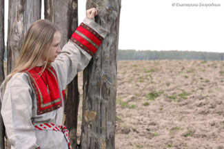 Vaaleahiuksinen nainen seisoo kyntämättömällä pellolla puuaidan vieressä pukeutuneena vanhanaikaiseen mekkoon.
