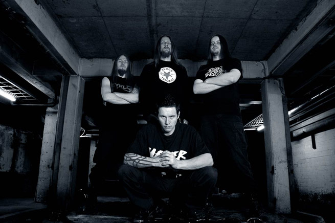 Kuvassa norjalainen extreme metal -bändi Zyklon. Yhtyeen kokoonpanoon kuului Samoth, Destructhor, Secthdamon ja Trym, jotka kaikki tulevat jatkamaan metallimusiikin luomista jatkossakin. Lisäksi ryhmän rumpali Trym ansaitsee elantonsa tatuointiliikkeensä pyörittämisessä bänditoiminnan ohella. Tämän kuvan ottaneen henkilön nimeä ei ole tiedossa. Harmaasävyinen valokuva nelipäisen Zyklonin kokoonpanosta. Kuvassa kolme miestä taustalla seisomassa ja etummaisena polvistuneena mies, jolla lyhyt musta tukka. Kaikki kuvan jäsenet ovat miehiä. Kolmella taaimmaisella pitkät heviletit.