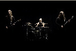Tummasävyinen promokuva kolmehenkisestä Chthonian-bändistä. Keskellä on rumpali rumpuineen — vasemmassa ja oikeassa laidassa kitaristi sekä basisti. Taustalla on pelkkää mustaa.