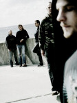 Soilwork-bändin jäsenet seisovat rannikolla jonkinlaisessa satamassa tai laiturilla. Etualalla oikeassa laidassa näkyy sumea sivuprofiili miehestä, jolla on viikset. Taustalla neljä miestä.