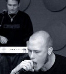 Mustavalkoinen valokuva Product Of Rage -yhtyeen laulajasta, joka etualalla pitää mikrofonia avoimen suun edessä, ja taaempana näkyvästä kosketinsoittajasta, jolla on musta paita ja kaulassa riipus tai koru.
