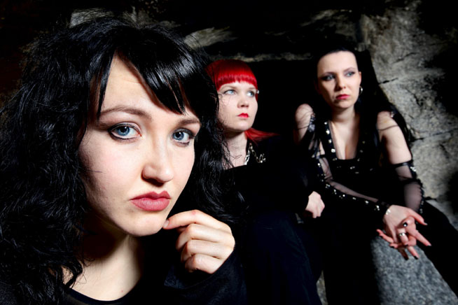 Ryhmäkuva kolmesta mustiin pukeutuneesta Kamala-bändin naisesta. Keskimmäisellä on punaiset hiukset, muilla mustat.