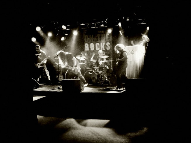 Harmaasävyinen ryhmäkuva Oceanwaken kokoonpanosta soittamassa On The Rocksin lavalla.