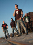 Volbeat-bändin jäsenet junaradan varressa. Taivas on syvän sininen ja pilvetön. Miehet pukeutuneet kesävaatteisiin.