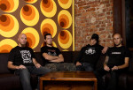 Rotten Sound -bändin jäsenet huoneen nurkkauksessa, jonka toinen seinä on paljasta tiiliseinää ja toisen seinän kuviona on kirkkaita kellertäviä ympyröitä. Kuvassa neljä miestä istuu pöydän ääressä.