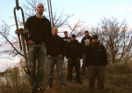 Total Devastation -bändin jäsenet, joita on kuusi, seisovat pöpelikössä. Taustalla taivasta ja lehdettömiä puita.