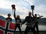 Gaia Epicus -bändin jäsenet seisovat rannalla. Kaksi miestä pitää kitaraa kohti taivaita ja keulahahmo heiluttaa Norjan lippua.